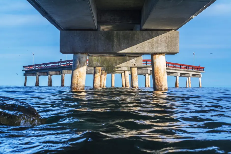 A view from beneath a concrete pier extending into the ocean on a clear day with calm water.